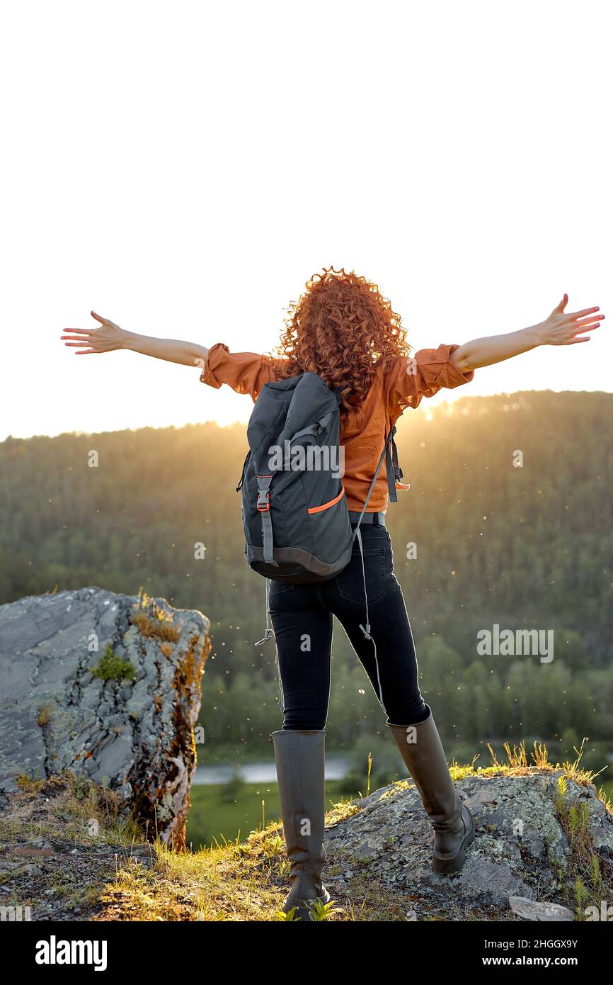 Vista posteriore su giovane donna caucasica snella con capelli ricci rossi che alzano le mani in montagna sensibilità libertà, al Sunshine del tramonto. Rilassato e libero, felice Foto Stock