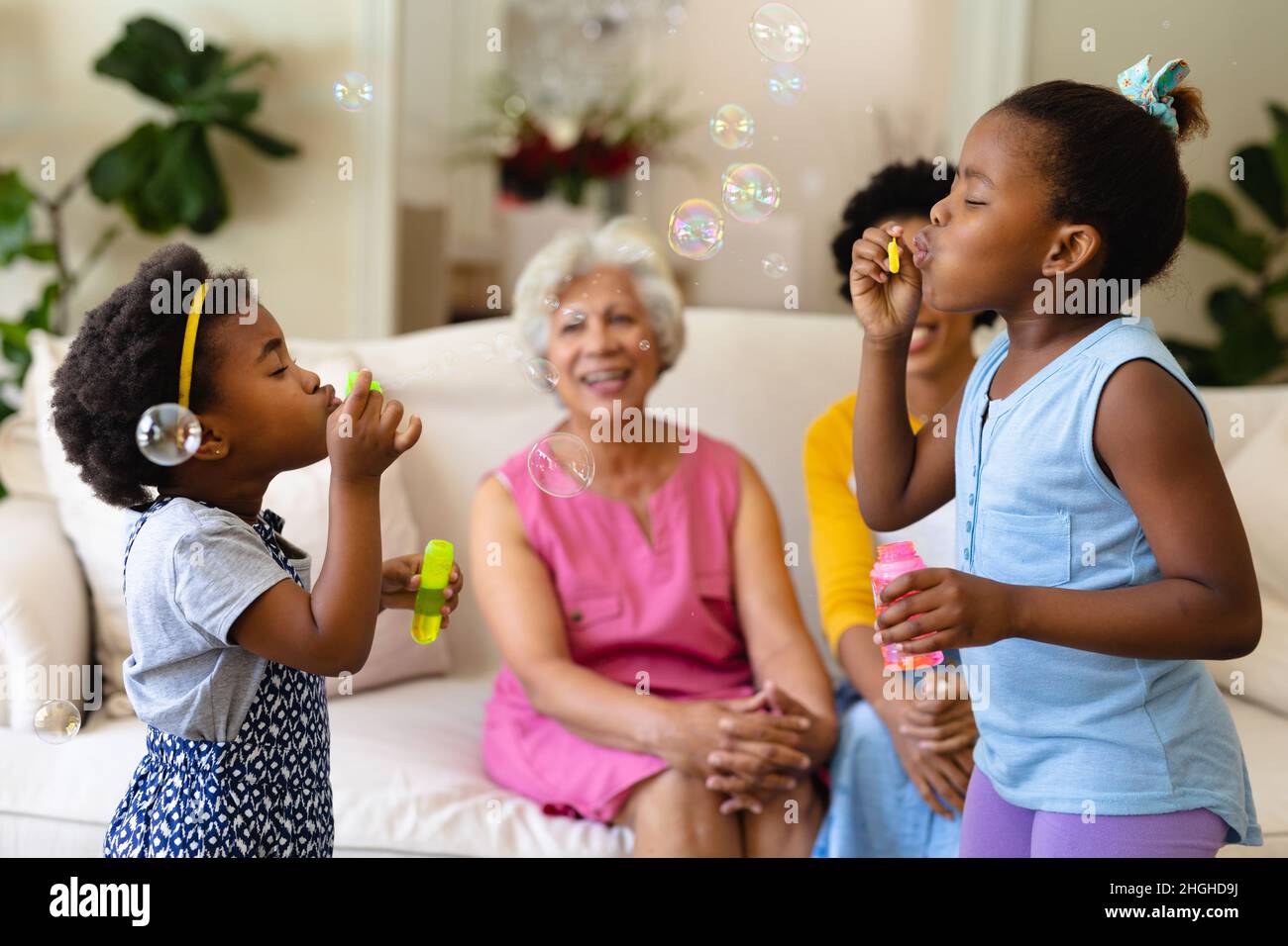 Due sorelle afroamericane che soffiano bolle mentre nonna e madre guardano seduti sul divano Foto Stock