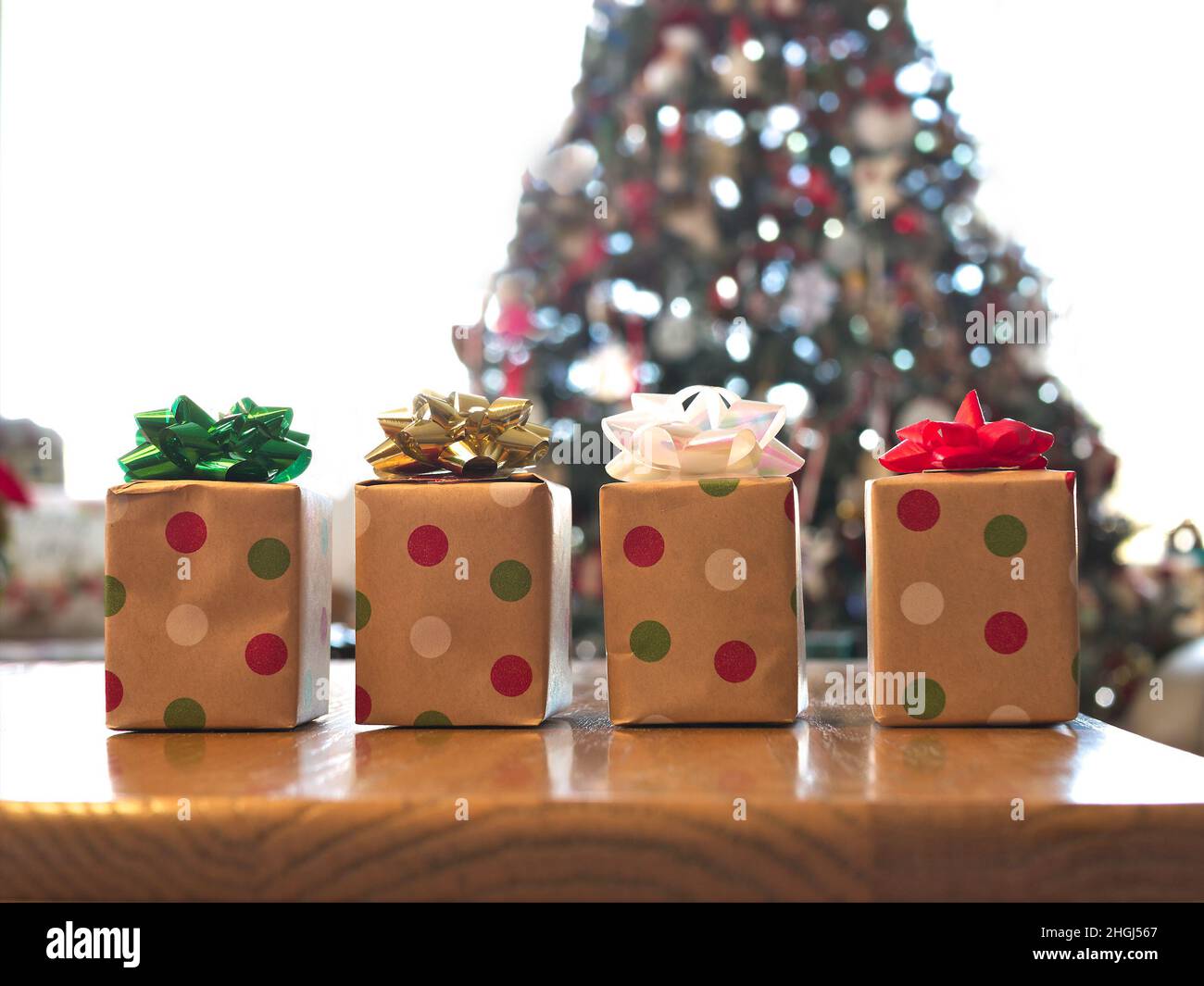Quattro scatole regalo splendidamente avvolte su tavola di legno con archi colorati, con albero di Natale decorato sullo sfondo. Foto Stock