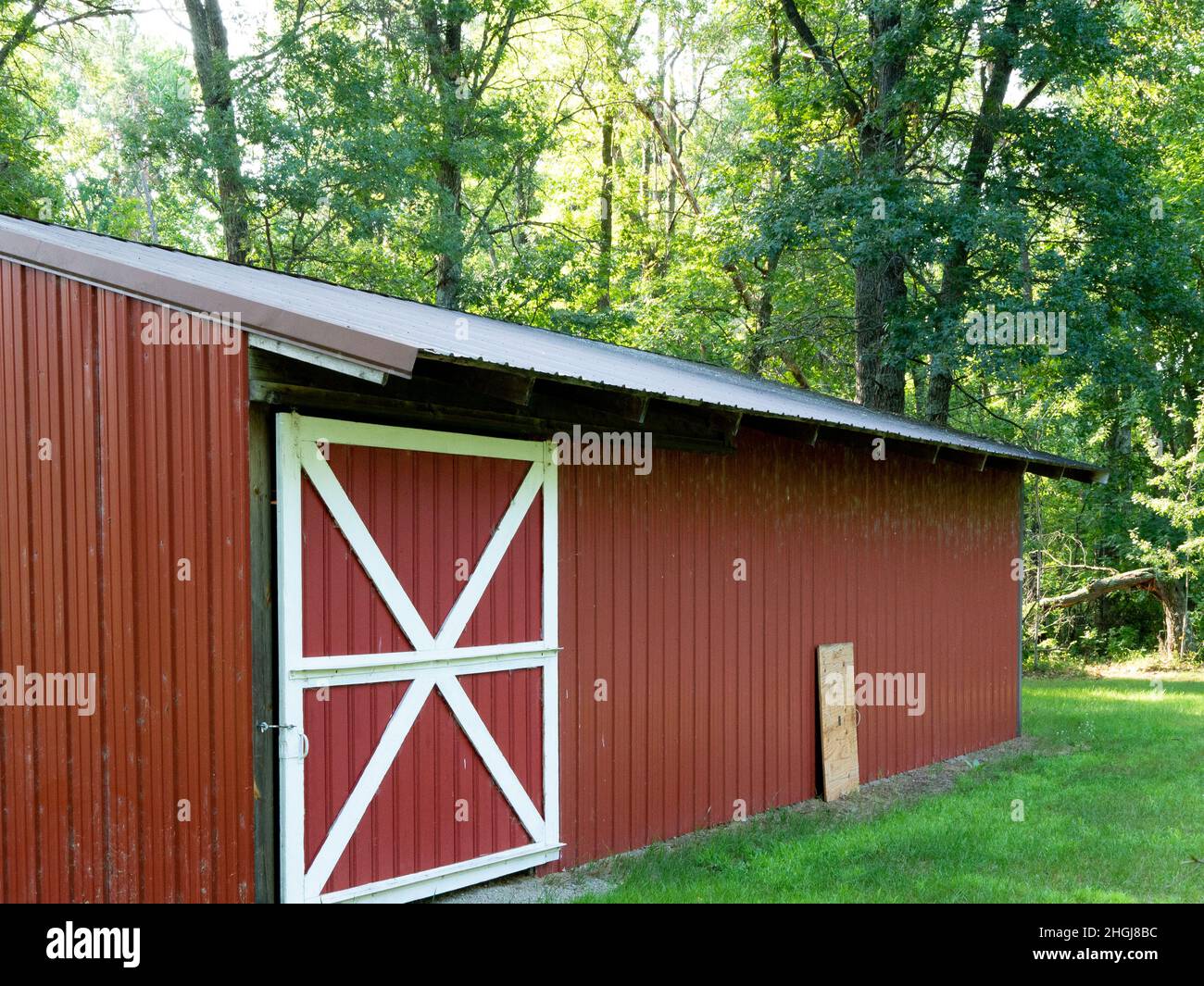 Il capannone di metallo rosso polveroso e sporco in azienda ha una porta scorrevole con rivestimento bianco. Circondato da erba verde e alberi. Foto Stock
