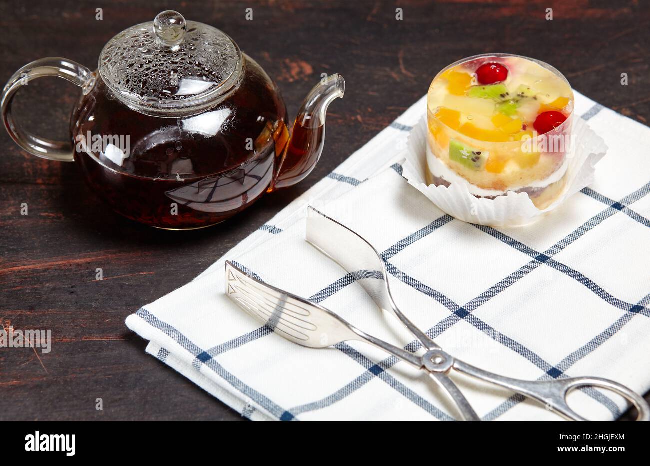 Tè nero in una teiera di vetro e torta con gelatina su sfondo di legno. Squisito mini dessert e tè tradizionale a colazione, primo piano Foto Stock