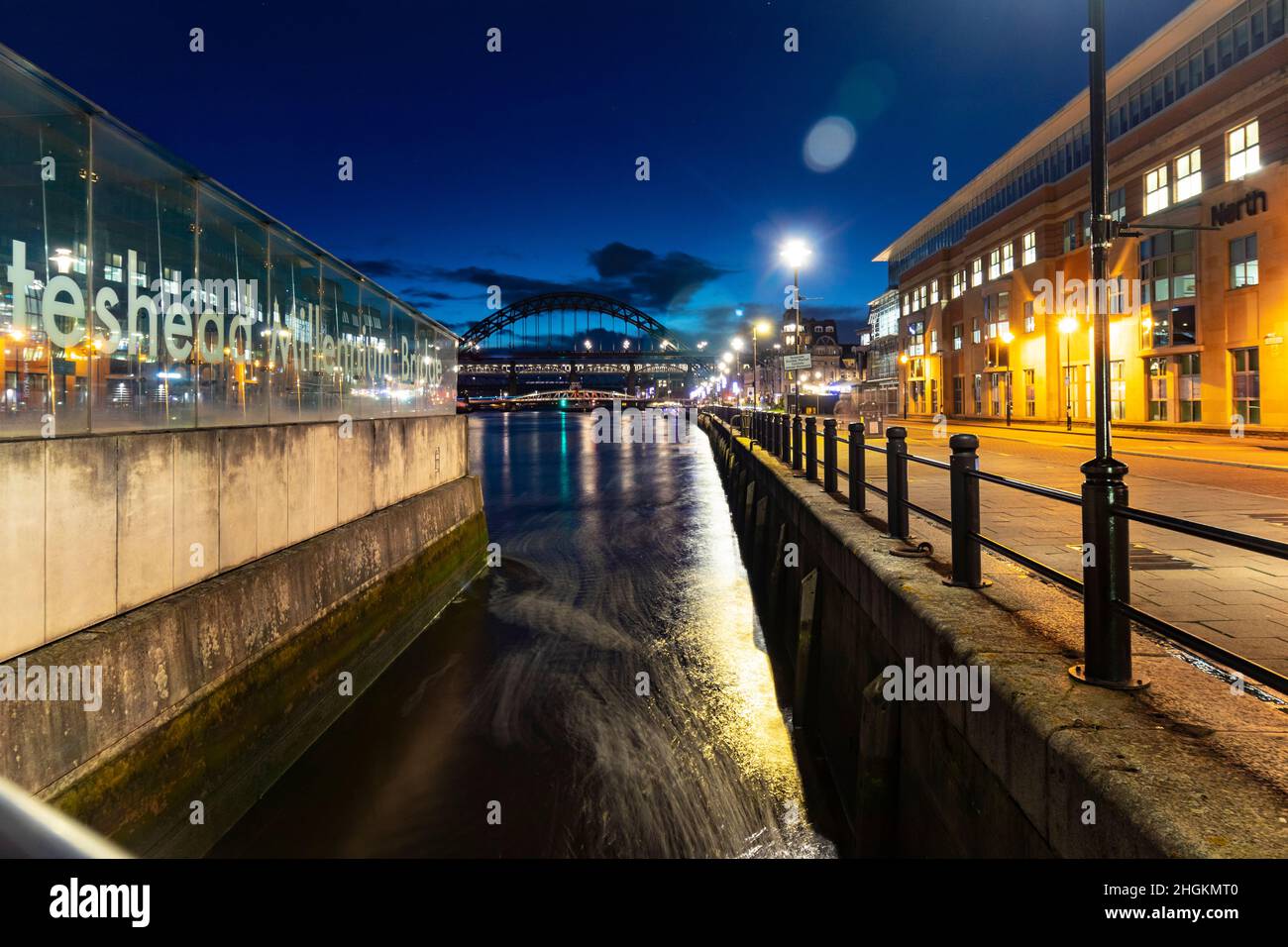 Vista notturna di un fiume con riflessi di luci della città, un edificio di vetro sulla sinistra e un ponte sullo sfondo. a newcastle upon tyne, ingresso Foto Stock
