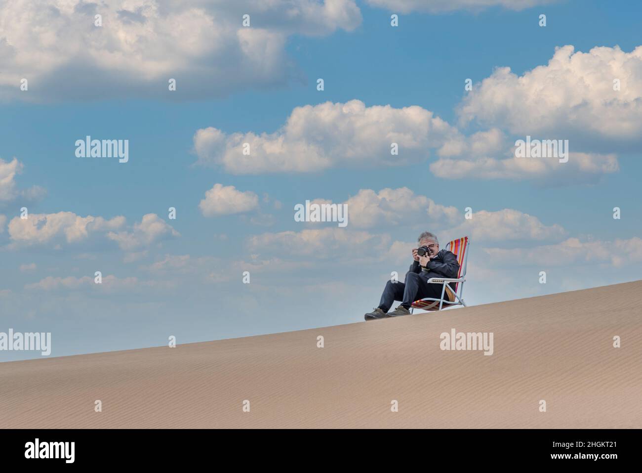 Vista di un maschio adulto seduto su una sedia da spiaggia nella parte superiore di una duna scattando una foto con la fotocamera. Foto Stock