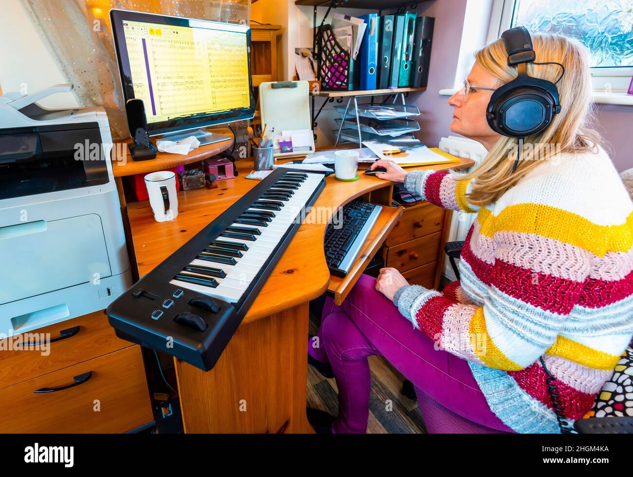 Musicista femminile, seduto alla sua scrivania di casa, di fronte a un PC monitor, utilizzando una tastiera controller midi, arrangiare una partitura musicale Foto Stock