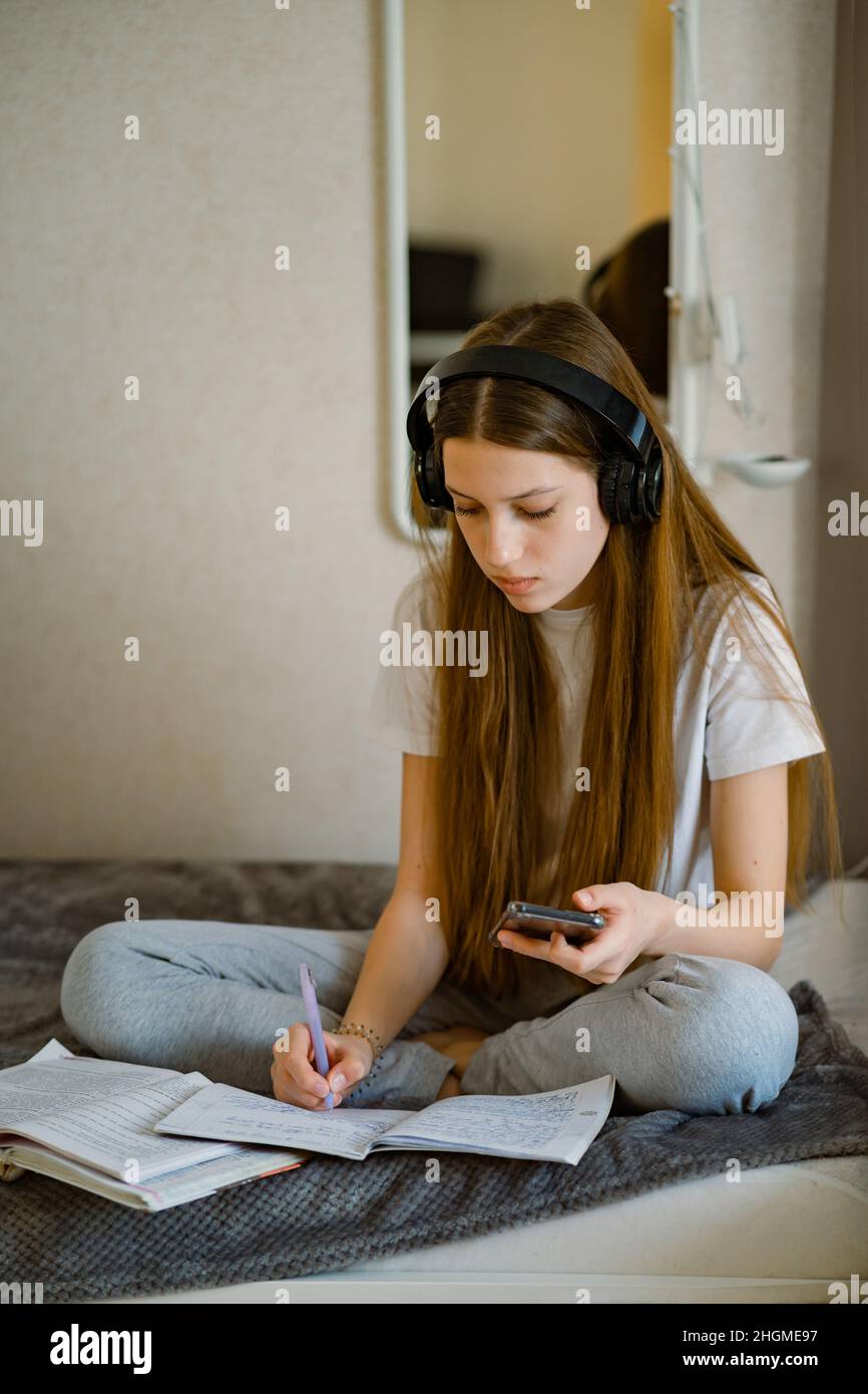 La ragazza adolescente a casa a letto in cuffie e con smartphone, prepara le lezioni. Foto Stock