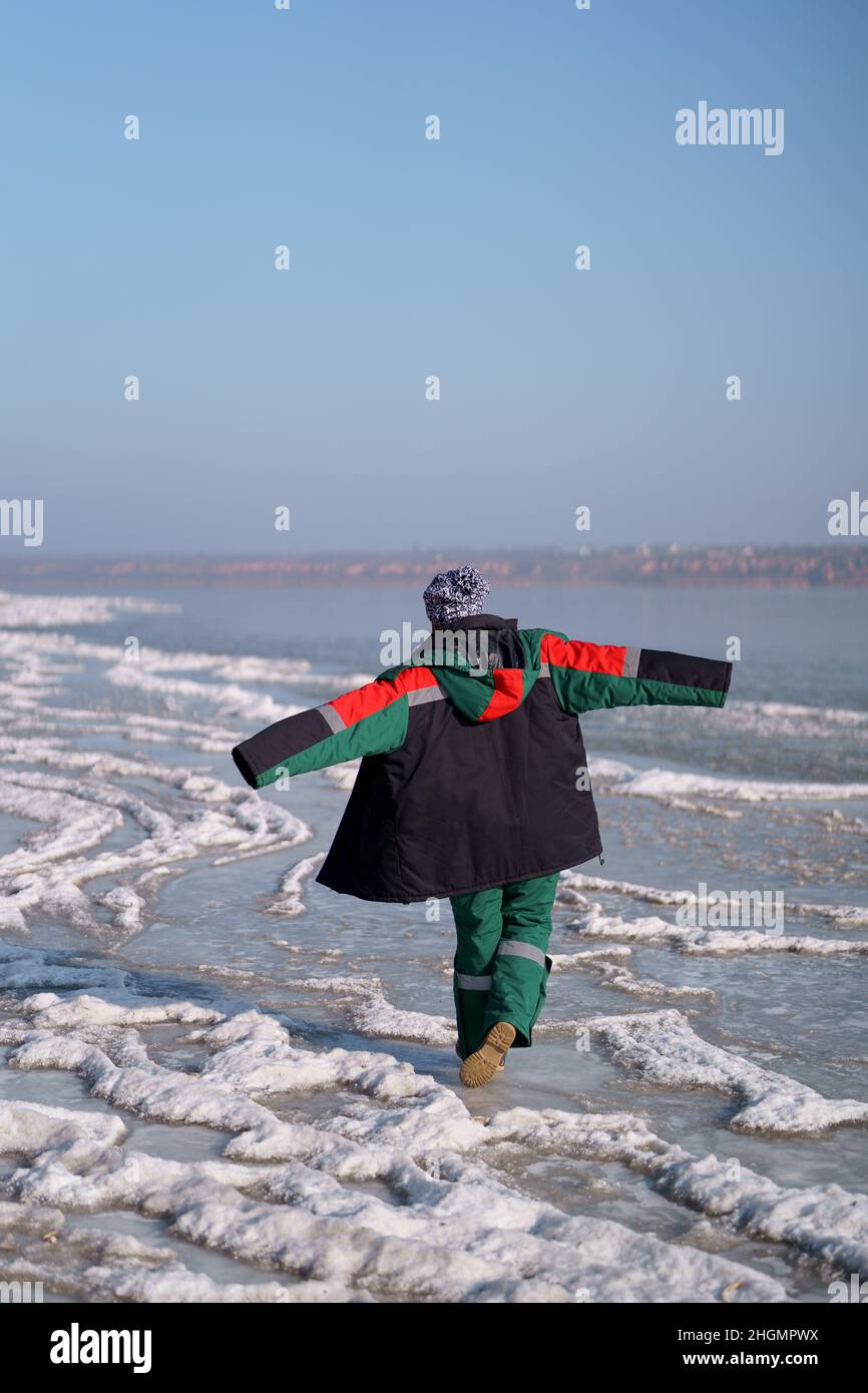 Una donna in un colorato costume termale che gode l'inverno mentre si erge su un lago ghiacciato con cielo blu sullo sfondo. Libertà Foto Stock