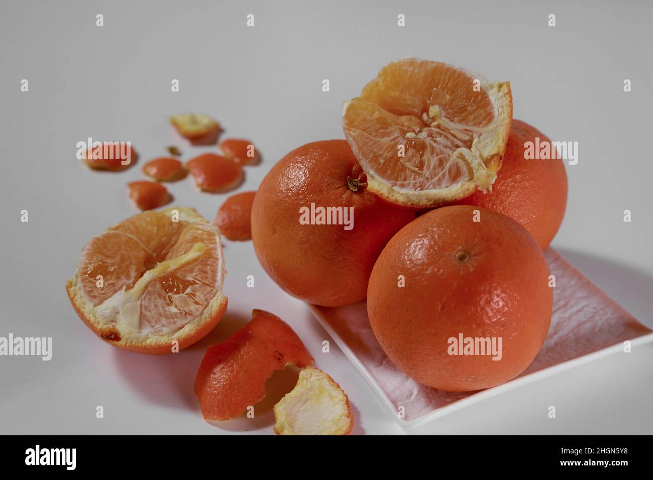 Quattro arance giacciono su una piastra di ceramica bianca e una è rotta a metà con una buccia sbucciata su uno sfondo bianco. Foto Stock