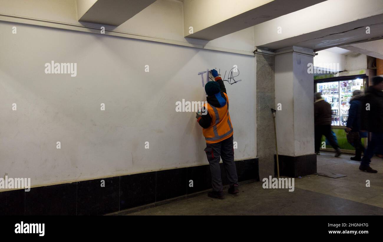 Rimozione dei graffiti dai vandali nel passaggio sotterraneo. Dipendente della città, indossando una uniforme arancione, pulisce le pareti dai graffiti e dalla scrittura, con un rosso Foto Stock