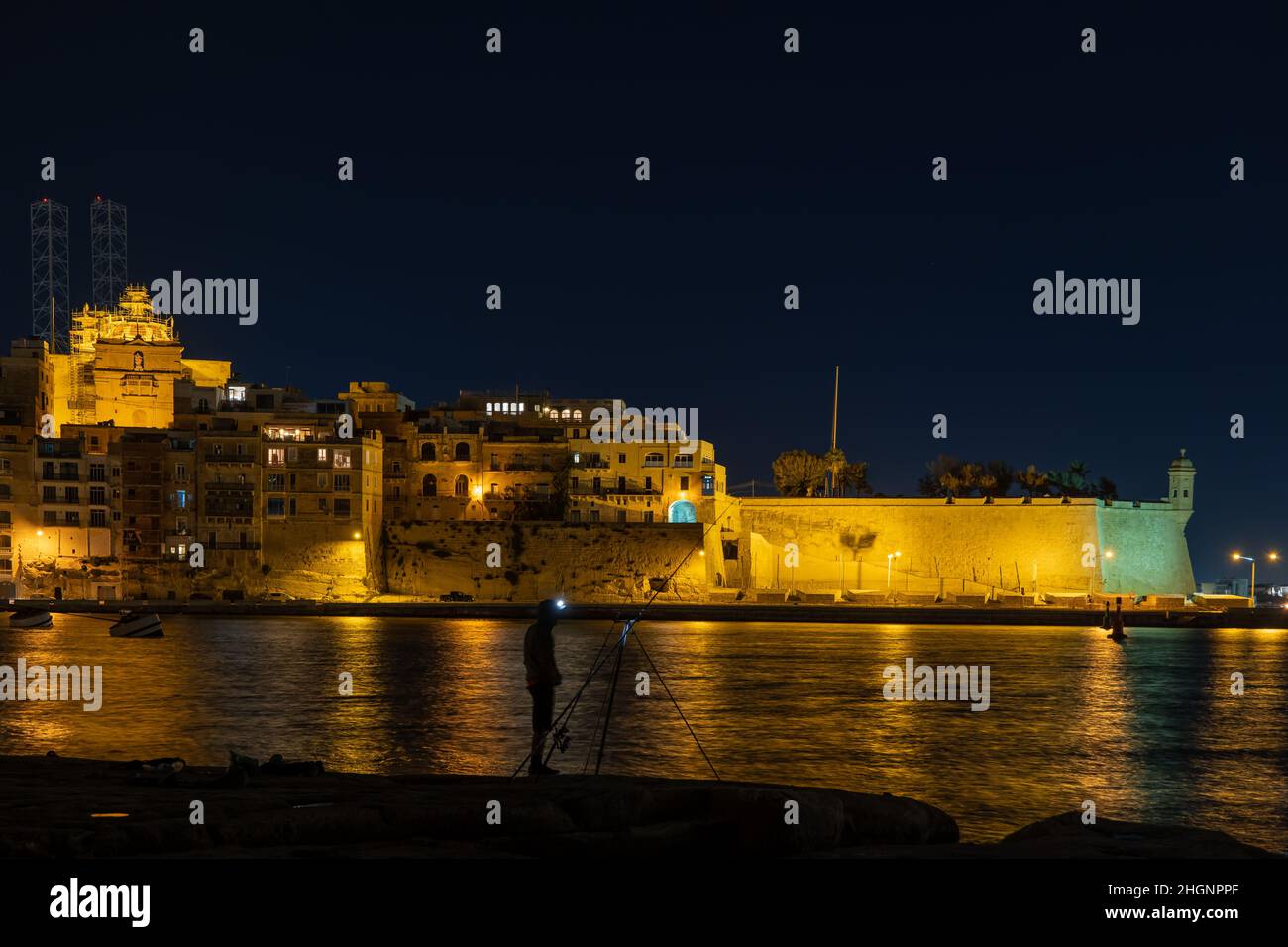 Skyline della città di Senglea (L-Isla, Civitas Invicta) di notte a Malta, solitario pescatore sulla riva del Grand Harbour. Foto Stock