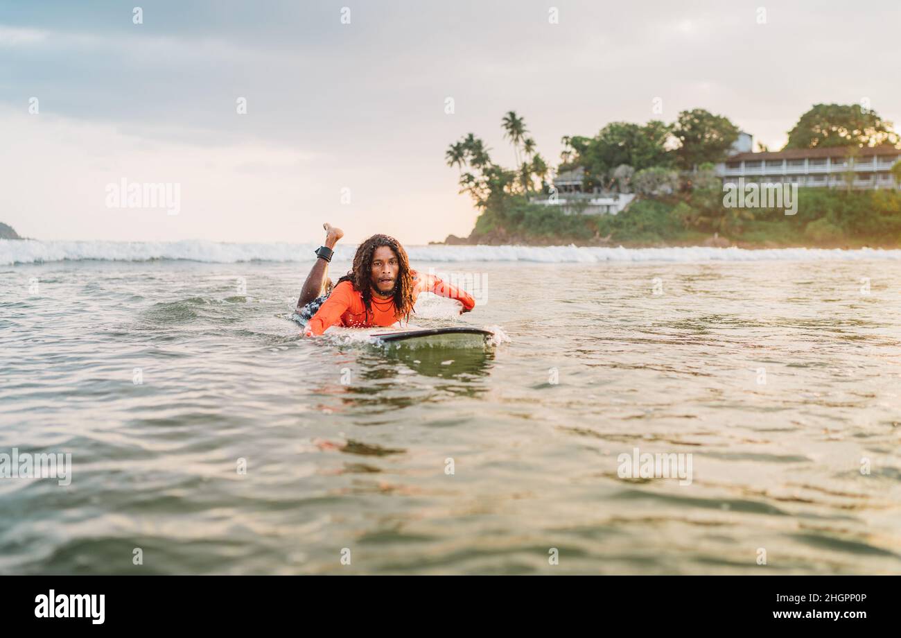 Uomo nero dai capelli lunghi che si paddling su una lunga tavola da surf al punto di surf nell'oceano Indiano. Palmeto ha littato raggi del tramonto sullo sfondo. Acqua estrema s Foto Stock