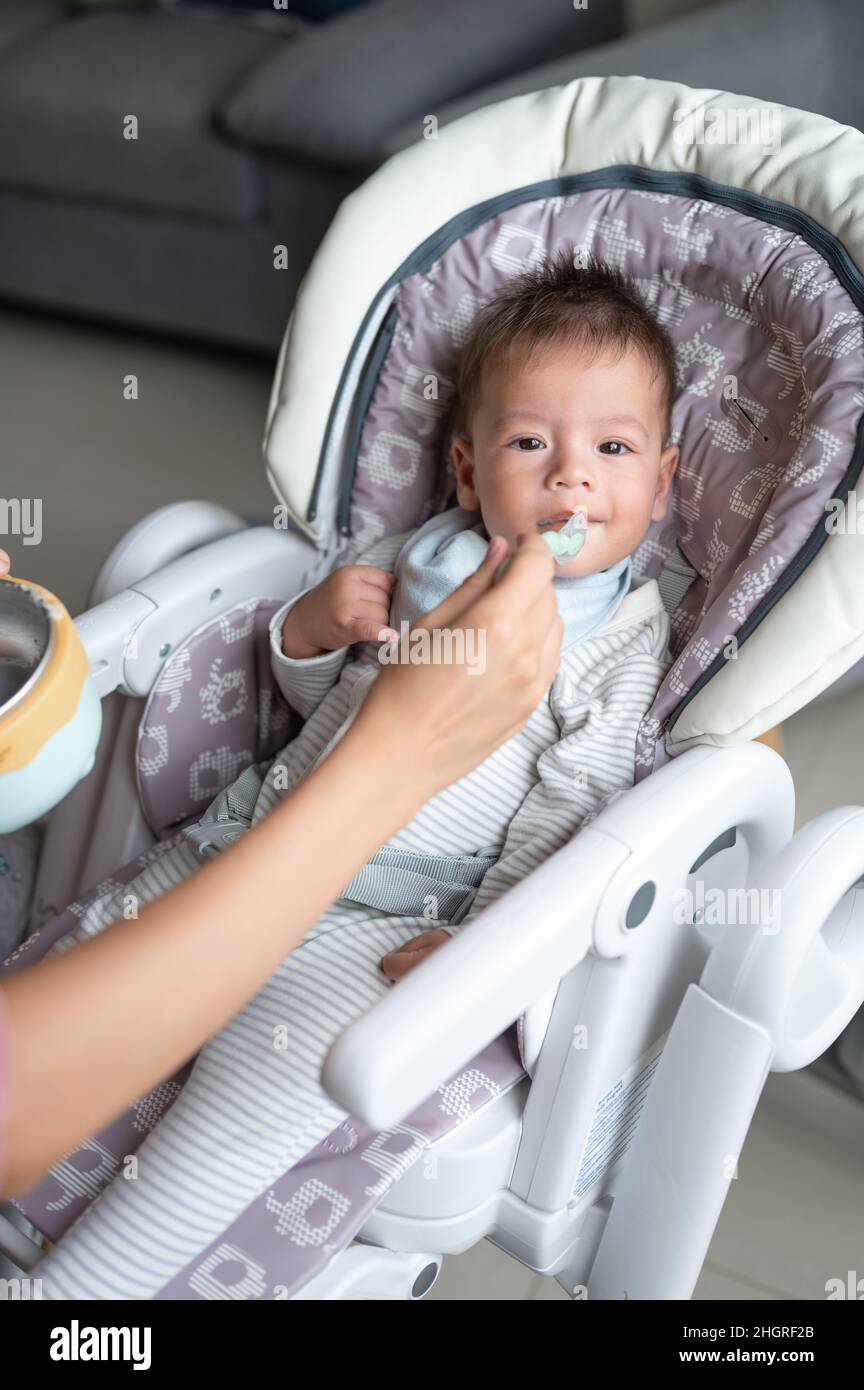 Madre che allatta il bambino con un cucchiaio in un seggiolone vicino alla finestra a casa Foto Stock