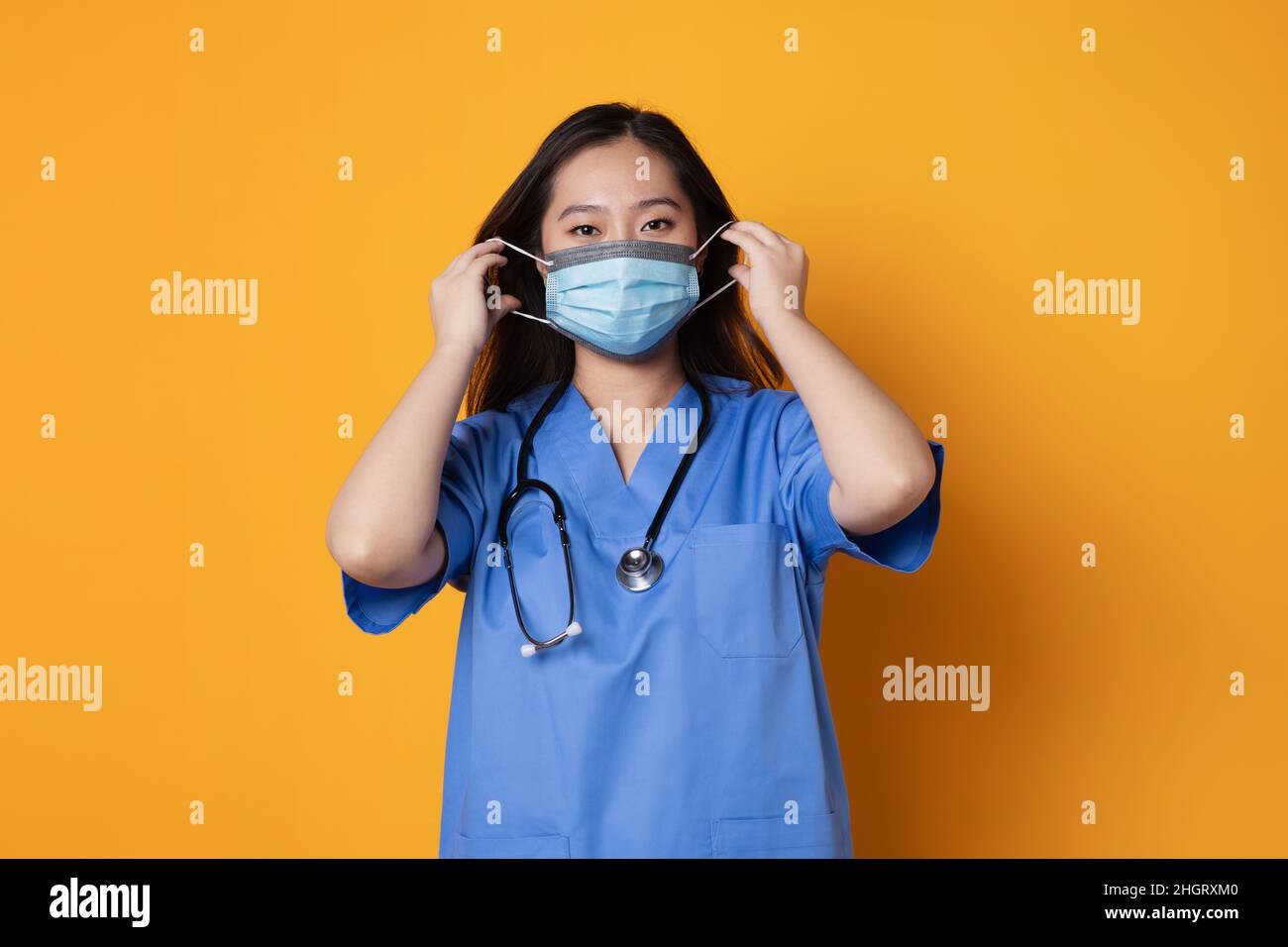 Foto studio con sfondo arancione di un medico asiatico che regola una maschera igienica sul suo viso Foto Stock