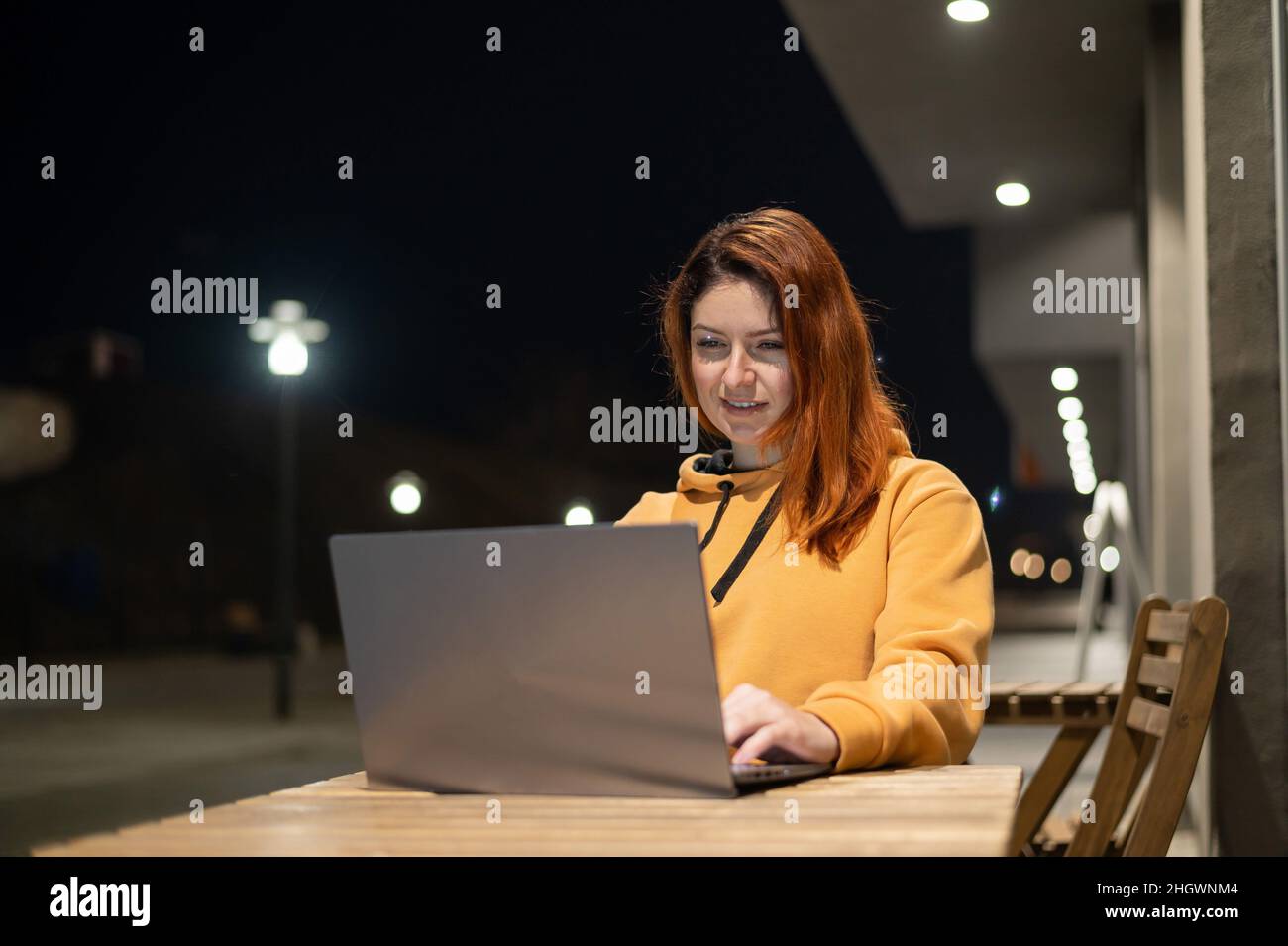 Una donna lavora a distanza a un computer portatile in un bar estivo a tarda sera. Ragazza felice studiare mentre si siede su una strada vuota ad un tavolo di legno Foto Stock