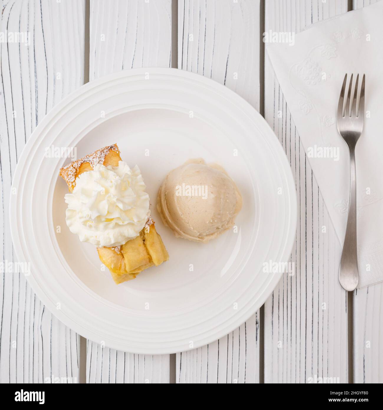 Pezzo di torta di mele con base di biscotto e panna montata, servito con gelato alla vaniglia in primo piano su un piatto su sfondo bianco di legno, quadrato Foto Stock