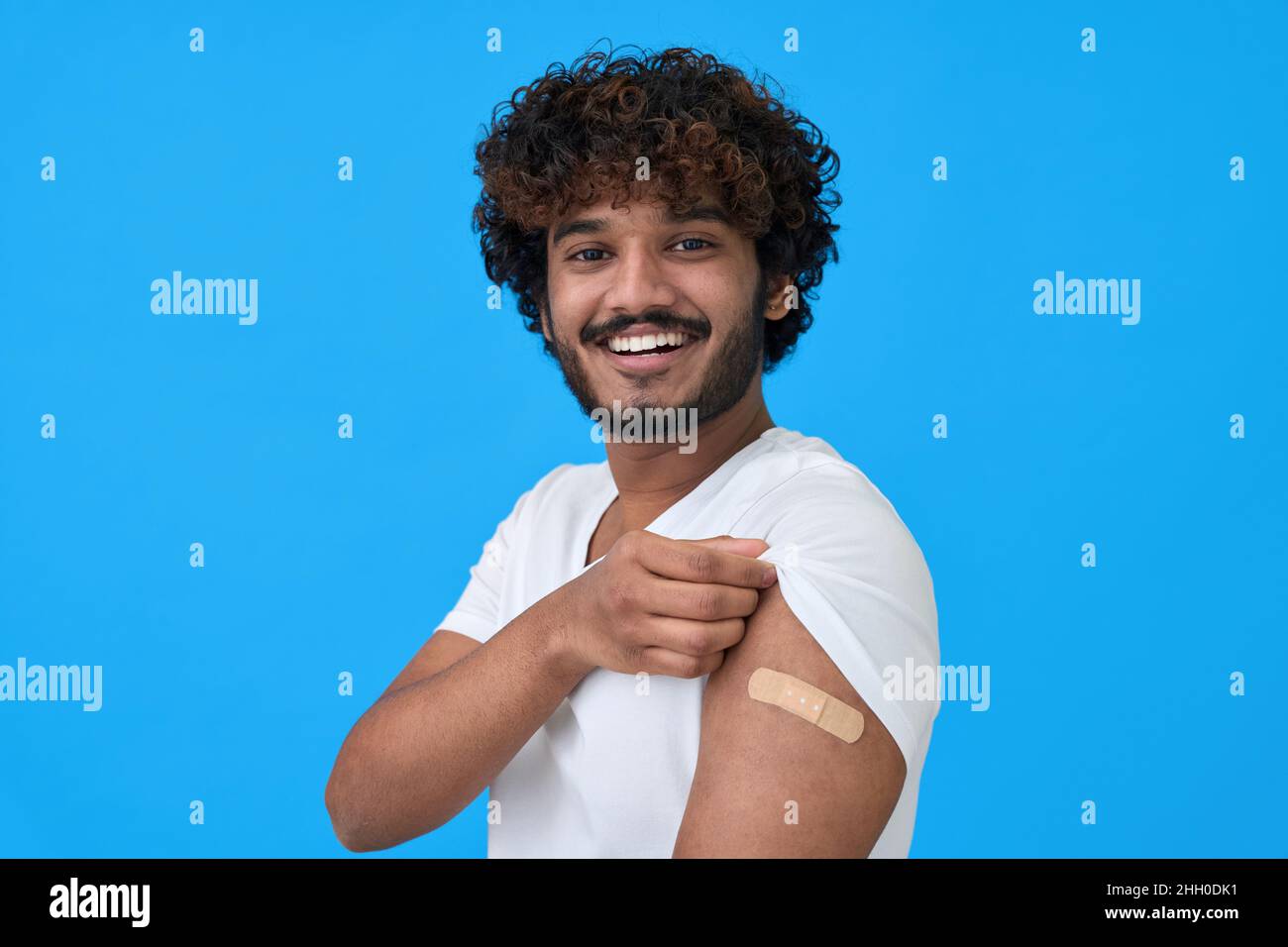 Felice giovane indiano che mostra bendaggio sul braccio dopo aver ottenuto la vaccinazione isolato su blu. Foto Stock