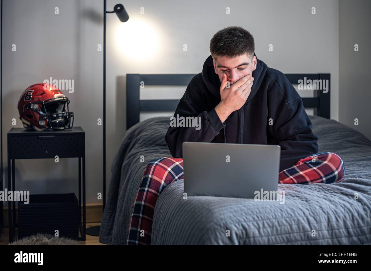 Un ragazzo si siede in una stanza su un letto e usa un computer portatile. Foto Stock