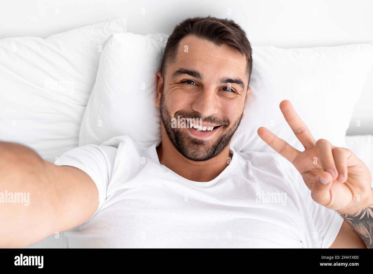 Sorridente millennial attraente blogger maschio con barba si sveglia sul letto bianco mostra segno di pace e fa selfie Foto Stock