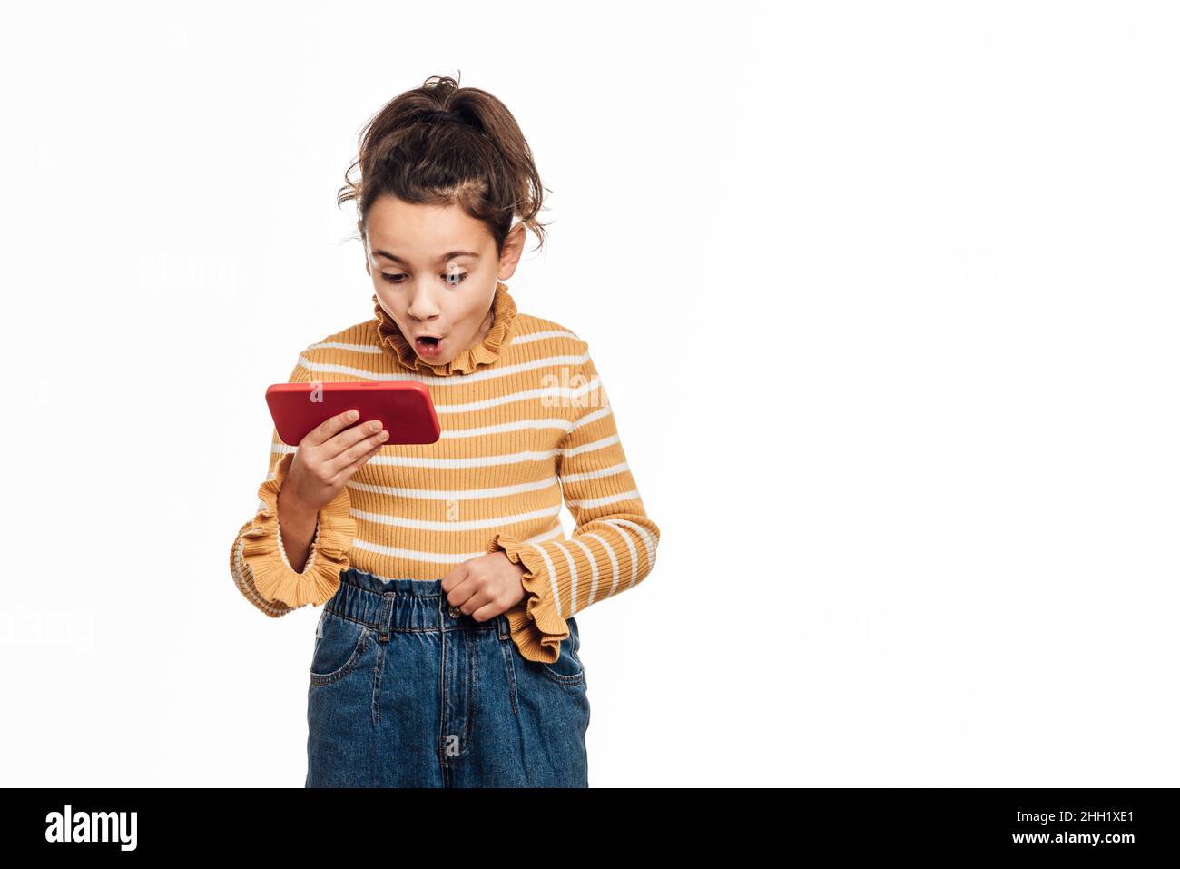 Bambina che guarda sorpreso lo schermo del telefono cellulare mentre si trova su uno sfondo isolato. Foto Stock