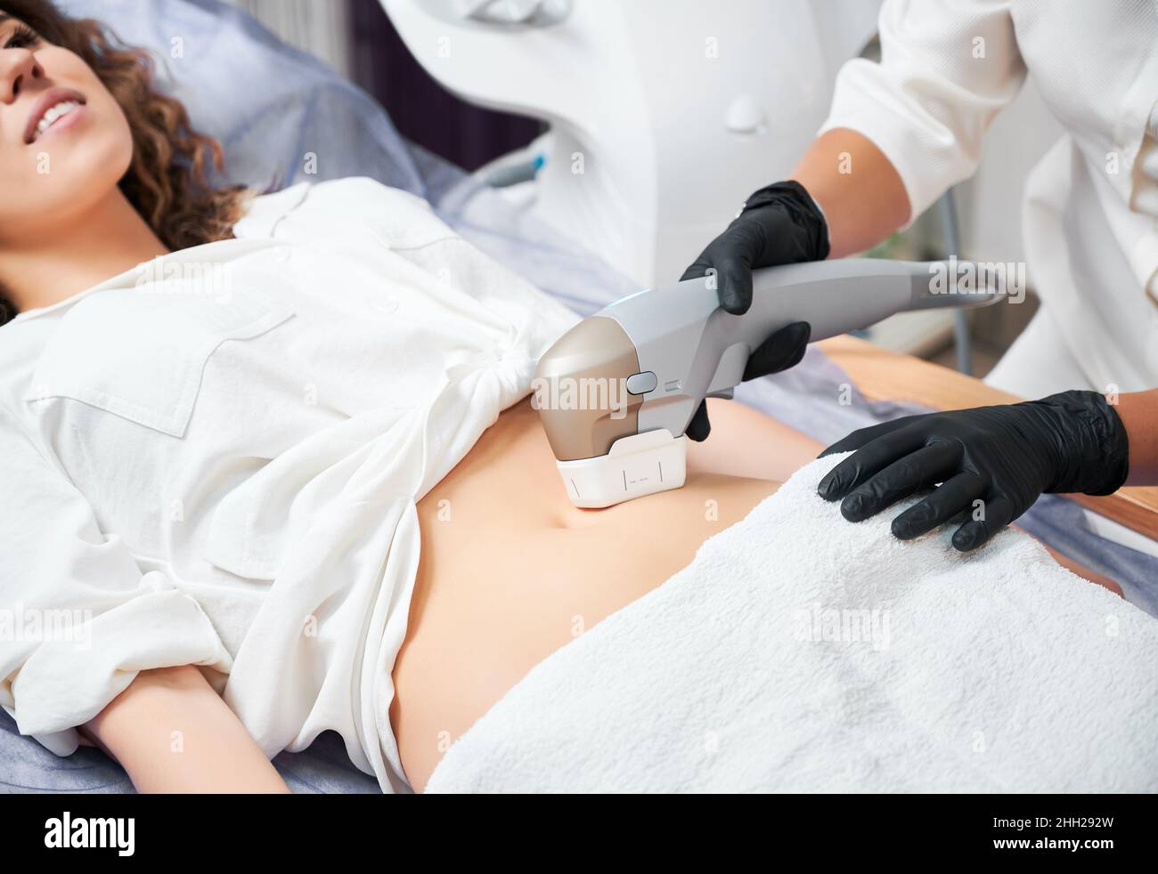 Primo piano di pazienti di sesso femminile che ricevono la procedura di irrigidimento e contornazione del corpo in clinica di cosmetologia. Estetista utilizzando un dispositivo a ultrasuoni durante l'esecuzione della procedura di sollevamento su addome donna. Foto Stock