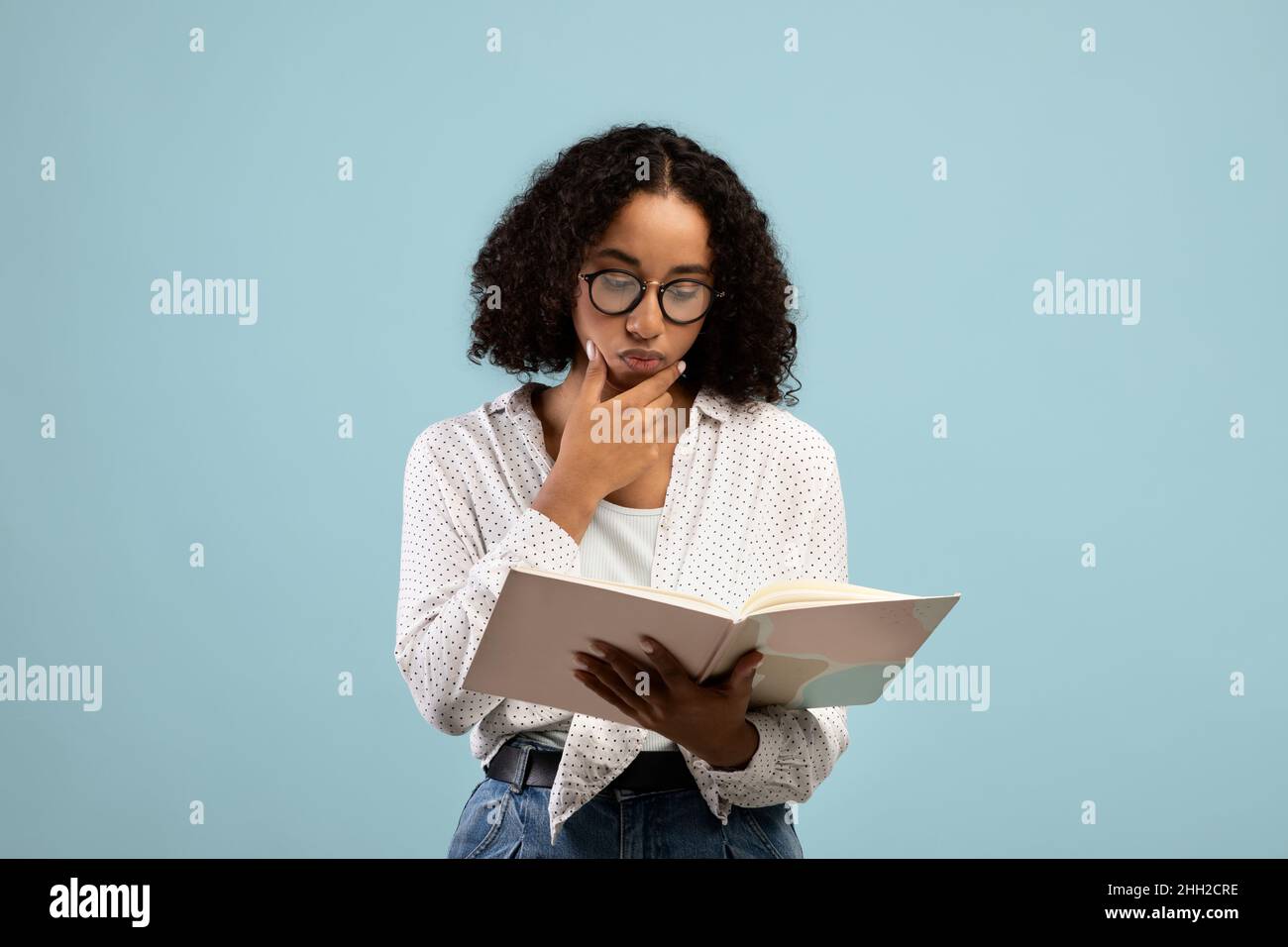 Studentessa femminile nera pensiva in occhiali lettura libro di testo, pensare sopra l'assegnazione, studiare per l'esame su sfondo blu Foto Stock