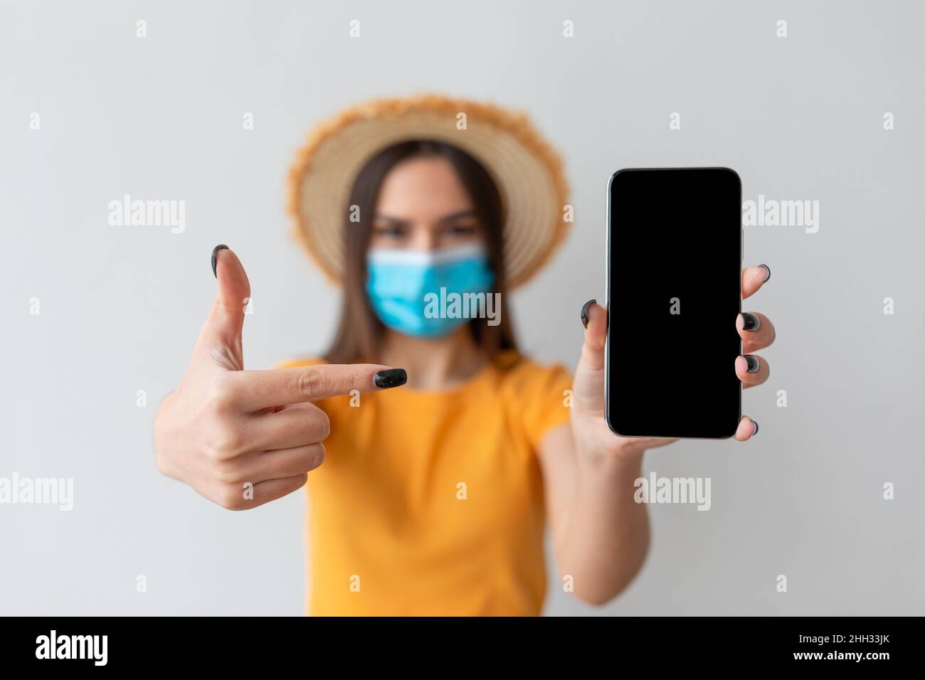 Giovane turista femminile in maschera medica che punta a smartphone con schermo vuoto, mockup per l'annuncio, app o sito web Foto Stock