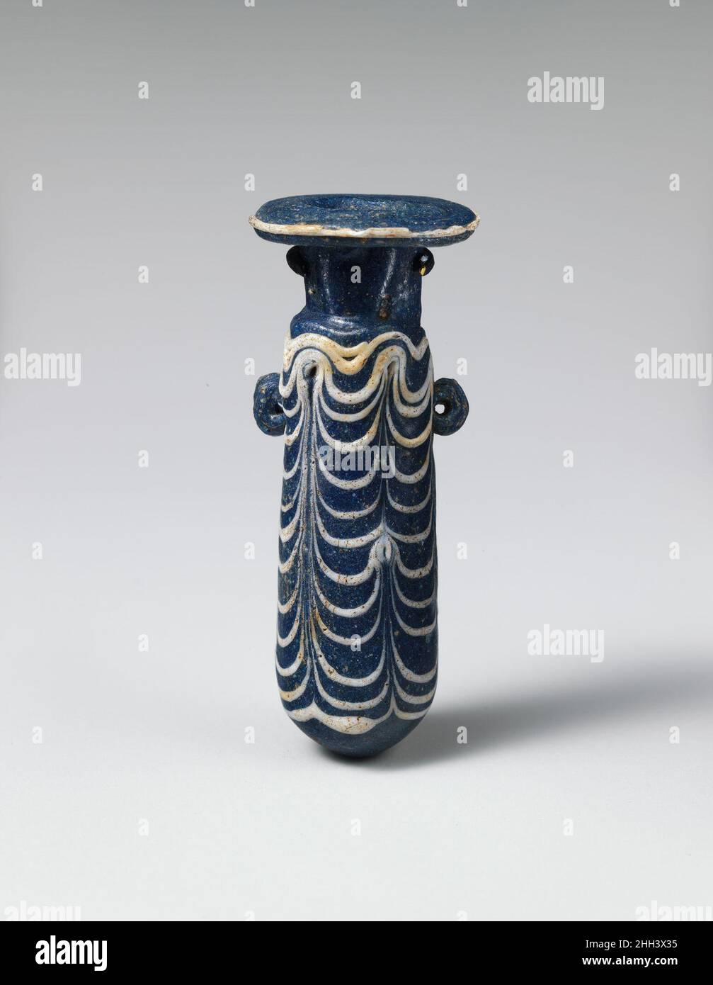 Alabastron di vetro (bottiglia di profumo) fine 4th–inizio 3rd secolo a.C. Greco, Mediterraneo orientale blu cobalto semi-opaco, con manici dello stesso colore; sentieri in bianco opaco.disco orizzontale ampio, realizzato come spirale attorno alla parte superiore del collo; collo cilindrico corto; spalla inclinata stretta; corpo cilindrico dritto; fondo convesso; Sul corpo superiore, due maniglie verticali dell'anello, forate, applicate sopra traccia pattern.A traccia attaccata a bordo di cerchio-disco; un'altra traccia applicata da spalla a bordo di fondo, tooled in un modello di festone con sei colpi verso l'alto, formando anelli a cima di body.intact, Foto Stock