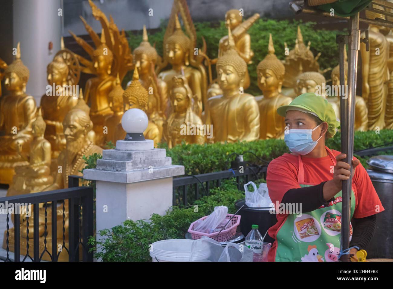 Un cuoco in una bancarella di cibo a Bangkok, Thailandia, guarda al suo fianco, un assortimento di statue di Buddha fatte in una fabbrica adiacente che fornisce un inusuale b/g Foto Stock
