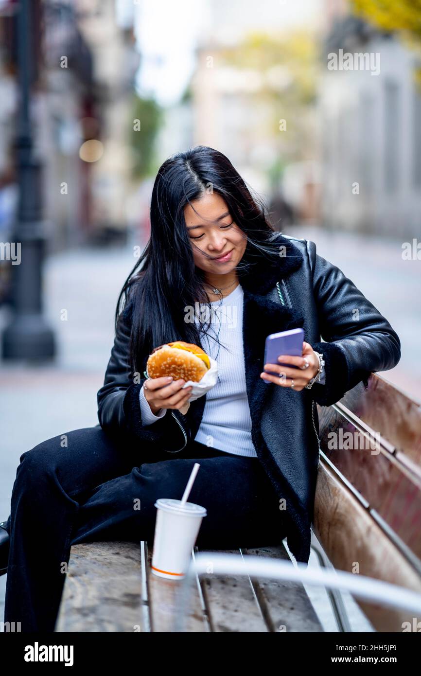 Donna con take out cibo utilizzando il telefono cellulare sulla panca Foto Stock
