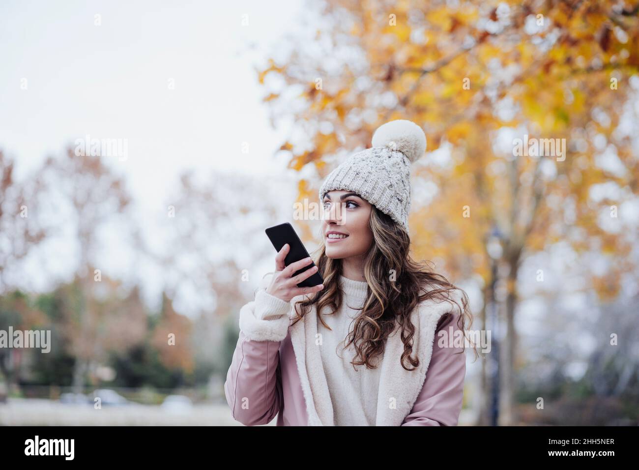 Donna con cappello a maglia che invia la posta vocale tramite telefono cellulare al parco Foto Stock