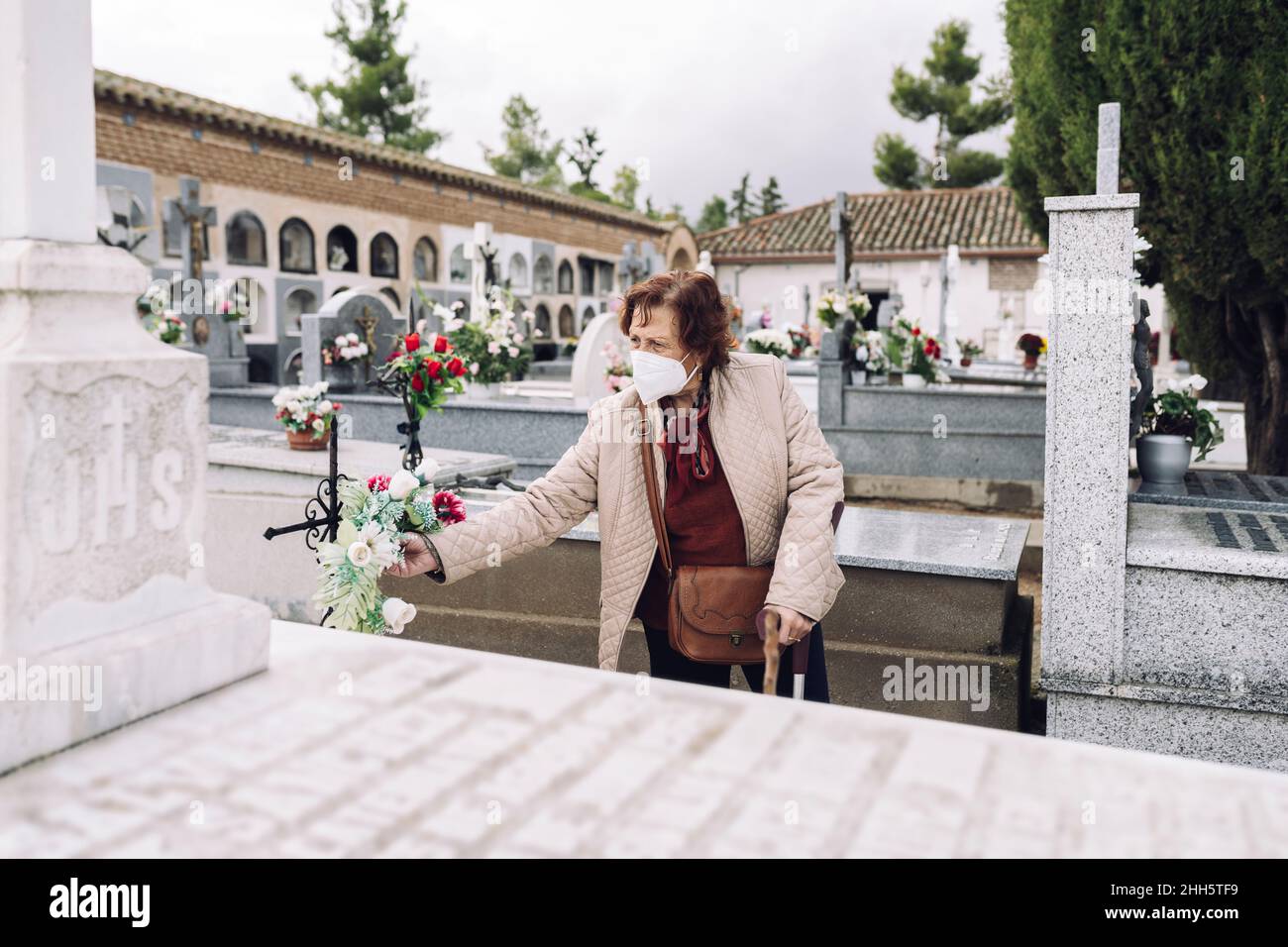 Donna anziana che indossa la maschera che mette i fiori sulla tomba nel cimitero Foto Stock