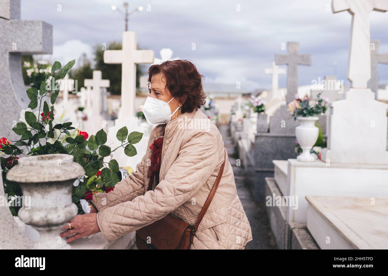 Donna anziana indossare maschera protettiva viso mettendo fiori sulla tomba nel cimitero Foto Stock