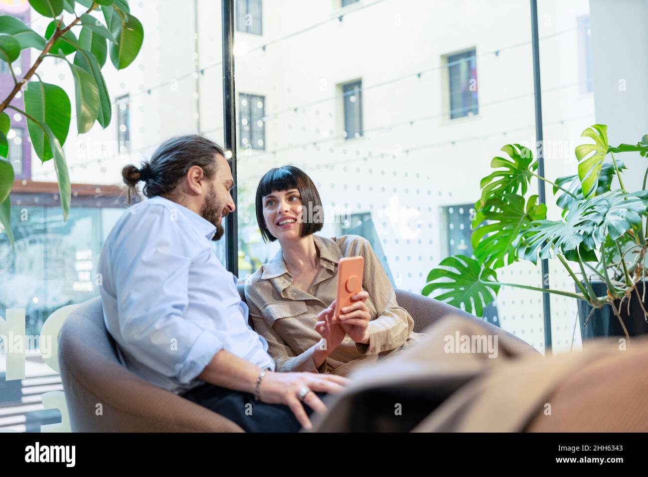 Donna sorridente che condivide lo smartphone con l'uomo seduto nella hall Foto Stock