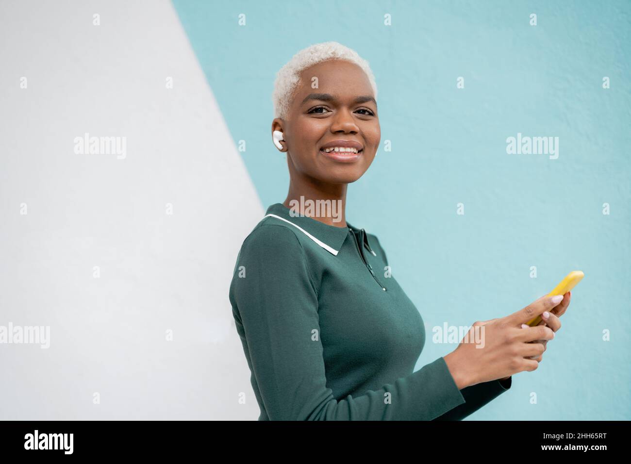 Donna sorridente che tiene lo smartphone indossando cuffie intrauricolari davanti alla parete Foto Stock