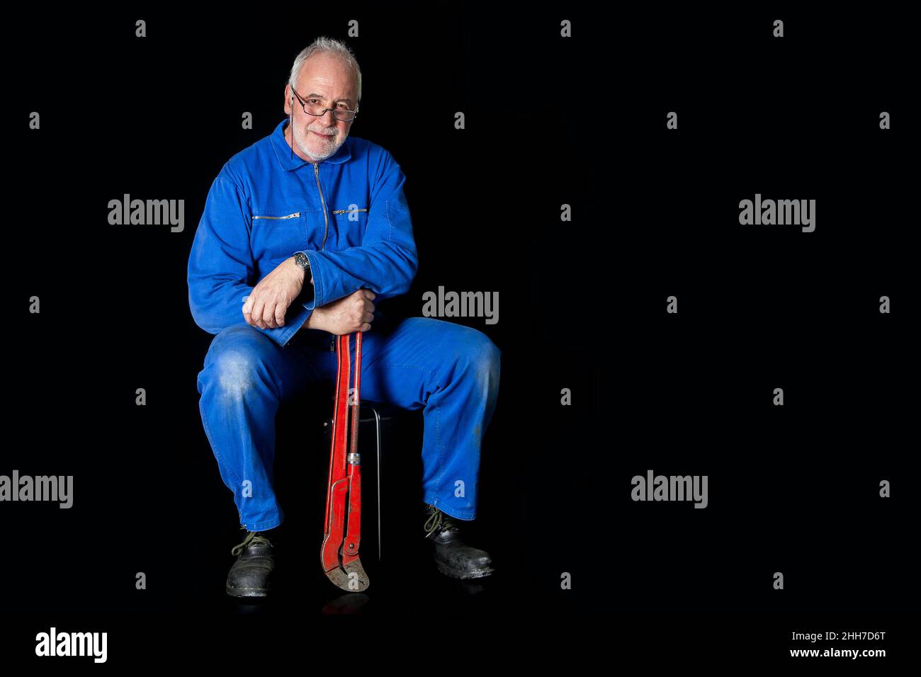 Un vecchio, esperto montatore in tuta blu si siede sulla sua custodia di montaggio su uno sfondo nero, tiene una grande chiave a tubo rossa nella sua mano. Foto Stock