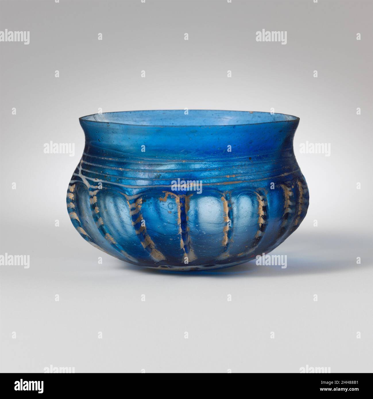 Coppa in vetro a coste metà 1st secolo d.C. blu traslucido romano; sentiero in bianco opaco.bordo svergolato, con bordo spaccato e molato; collo concavo corto; corpo globulare curvato verso il basso convesso poco profondo.Sentiero applicato come un grande blob e avvolto a spirale sul fondo e poi su lato al collo; Lato tooled in diciassette irregolari, verticali o slanting ribbs.intact, ma parti di sentiero mancanti attraverso l'invecchiamento; molte bolle, alcune grandi; vaiolatura di bolle di superficie, dulling, iridescence e le zone di tempo cremoso, particolarmente intorno costs.Bright ciotola blu con decorazione bianca del filetto, costola verticale Foto Stock