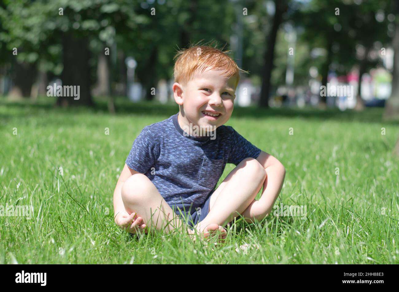 Un ragazzino dai capelli rossi siede nel parco sul prato e sorride. Il bambino siede nel sole diurno luminoso Foto Stock