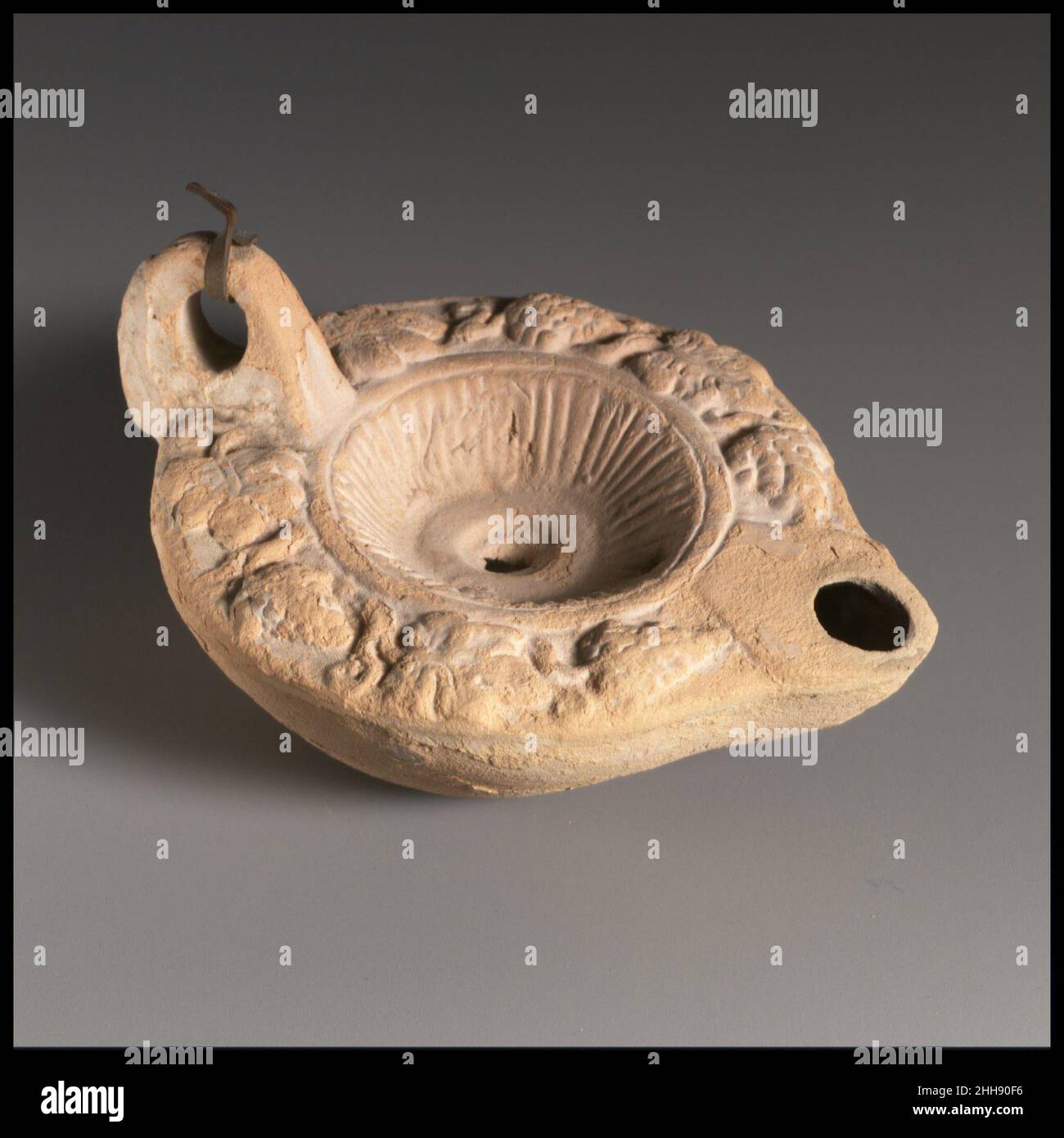 Lampada ad olio di terracotta 3rd–4th secolo d.C. Vessberg romano tipo 18a. Impugnatura ad anello, con stelo scanalato. Realizzato in stampo. Disco affondato con foro di riempimento centrale e linee radianti ravvicinate intorno al lato; foro più piccolo vicino al bordo del disco dietro l'ugello. A spalla in rilievo: Alternanza di foglie di vite e grappoli di uva. Su base piatta all'interno di un cerchio inscritto, in lettere greche incise: ΠΛΑ/TWN/OC.parte superiore del manico rotta e riparata. Buccia argilla.. Lampada ad olio di terracotta 241804 Foto Stock