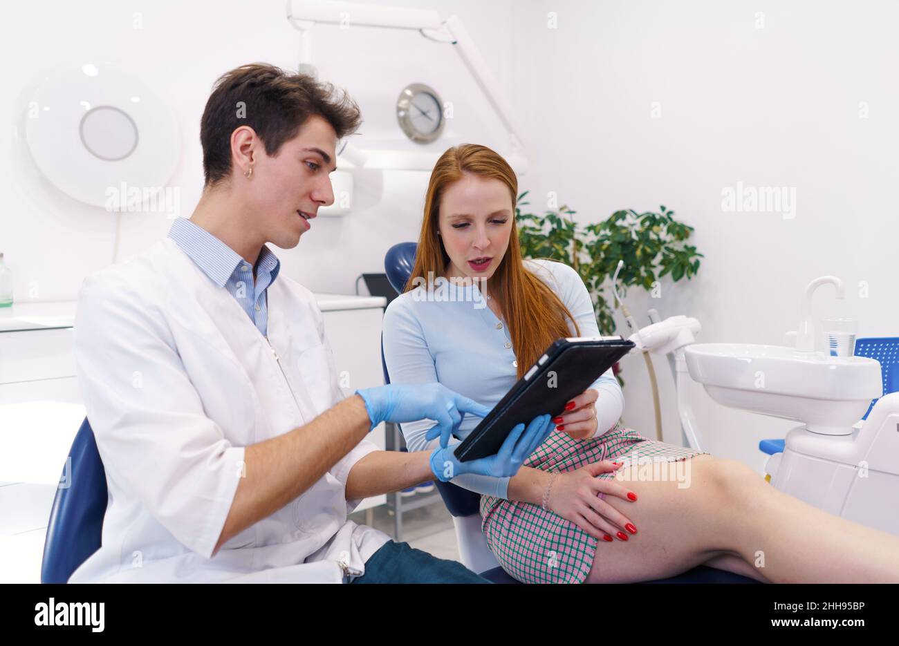 Giovane uomo in accappatoio bianco e guanti in lattice che mostrano i dati su tablet moderno a donna rossa mentre lavora in un moderno studio dentistico Foto Stock