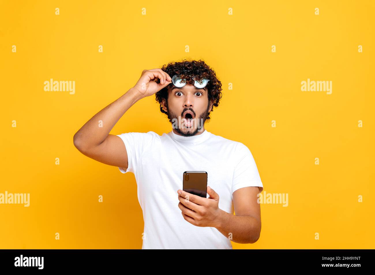 Sorpreso scioccato indiano ragazzo in t-shirt bianca, tiene uno smartphone in mano, toglie i suoi occhiali e guarda con stupore la fotocamera, ha ricevuto un messaggio inaspettato o notizie, isolato sfondo arancione Foto Stock