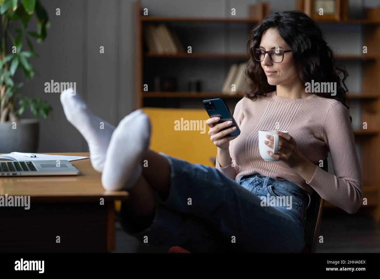 Ragazza rilassata freelance che riposa alla scrivania con telefono cellulare e tazza di caffè, godendo freelance Foto Stock
