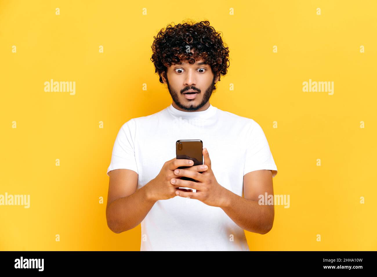 Sorpreso scioccato indiano ragazzo in t-shirt bianca, utilizza il suo smartphone, guarda in stupore lo schermo, ha ricevuto un messaggio inaspettato o notizie, stand su isolato sfondo arancione, espressione del viso perplesso Foto Stock