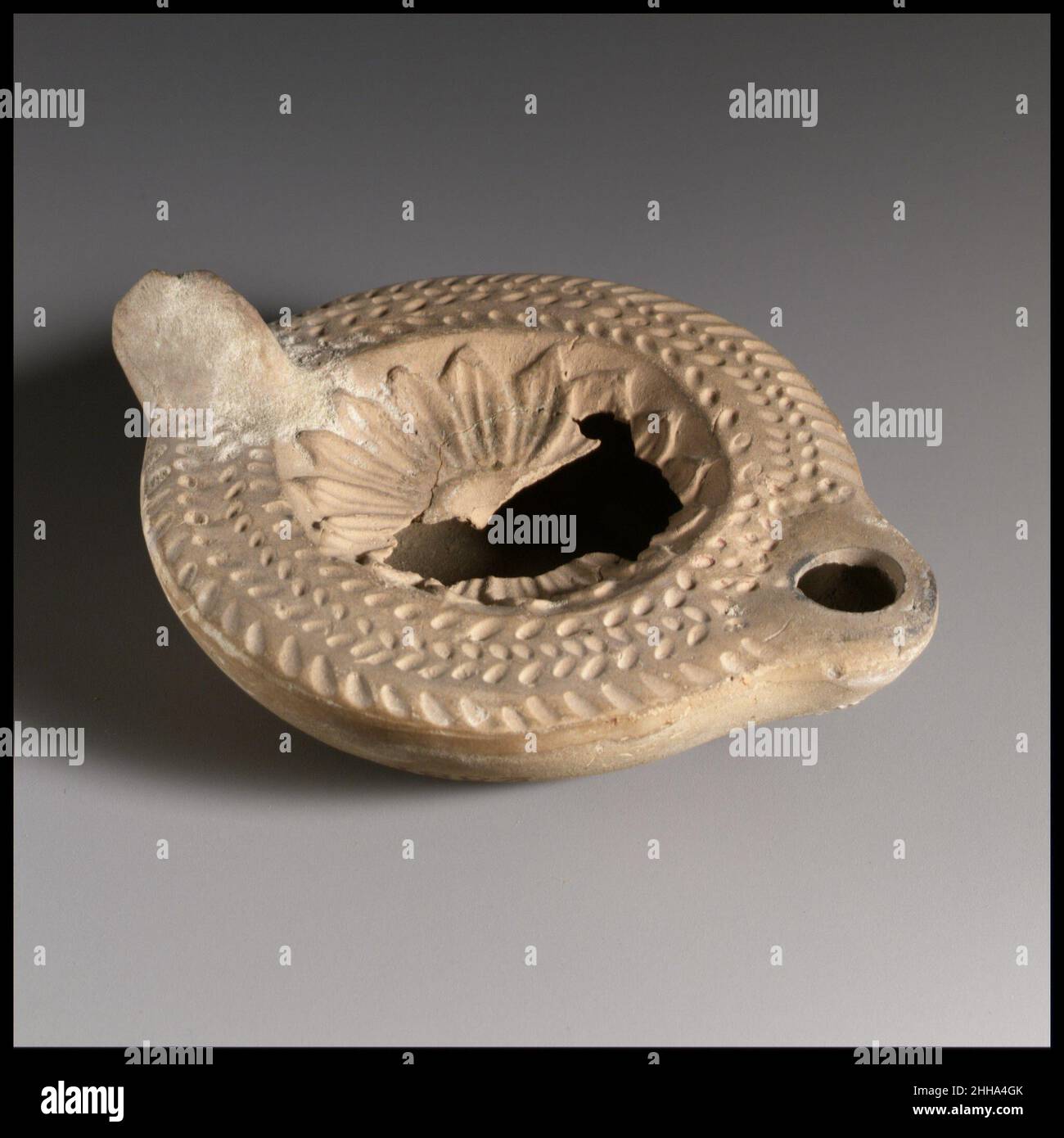 Lampada ad olio di terracotta fine 2nd–3rd secolo d.C. Loeschcke romano tipo 8. Impugnatura non forata. Realizzato in stampo. Discus: Rosetta di petali appuntiti sovrapposti. Su spalla larga quasi orizzontale: Due file di puntini impressi circondate da una corona a spina di pesce. Ugello arrotondato con grande foro per stoppino. Base concava non definita; sulla base, in caratteri greci: ?PMIANO. Buff creta. Parte di disco rotto e mancante, e un piccolo buco in lato a destra verso maniglia. Qualche incrostazione limosa sopra e intorno all'impugnatura. Lampada ad olio di terracotta. Romano. Fine 2nd–3rd secolo d.C.. Terracotta, modellata. Tardo imperiale. Terracotte Foto Stock