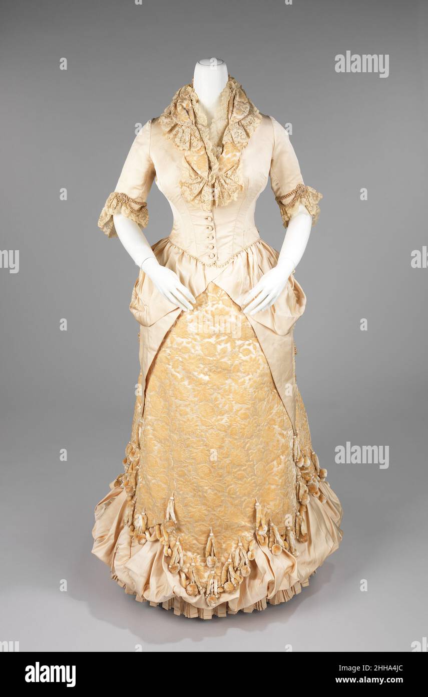 Abito da sera 1885 americano la silhouette del trambusto, anche se associata principalmente alla seconda metà del 19th secolo, ha avuto origine in prima moda come un semplice urto sul retro del vestito, come con fine 17th-inizio 18th secolo mantuas e fine 18th-inizio 19th secolo abiti Impero. La silhouette piena di trambusto ebbe il suo primo aspetto vittoriano alla fine del 1860s, che iniziò come pienezza nelle gonne che si spostavano verso la parte posteriore del vestito. Questa pienezza è stata elaborata in legami per camminare che ha creato un puff alla moda. Questo puff trendsetting si espanse ed è stato poi costruito con supporti Foto Stock