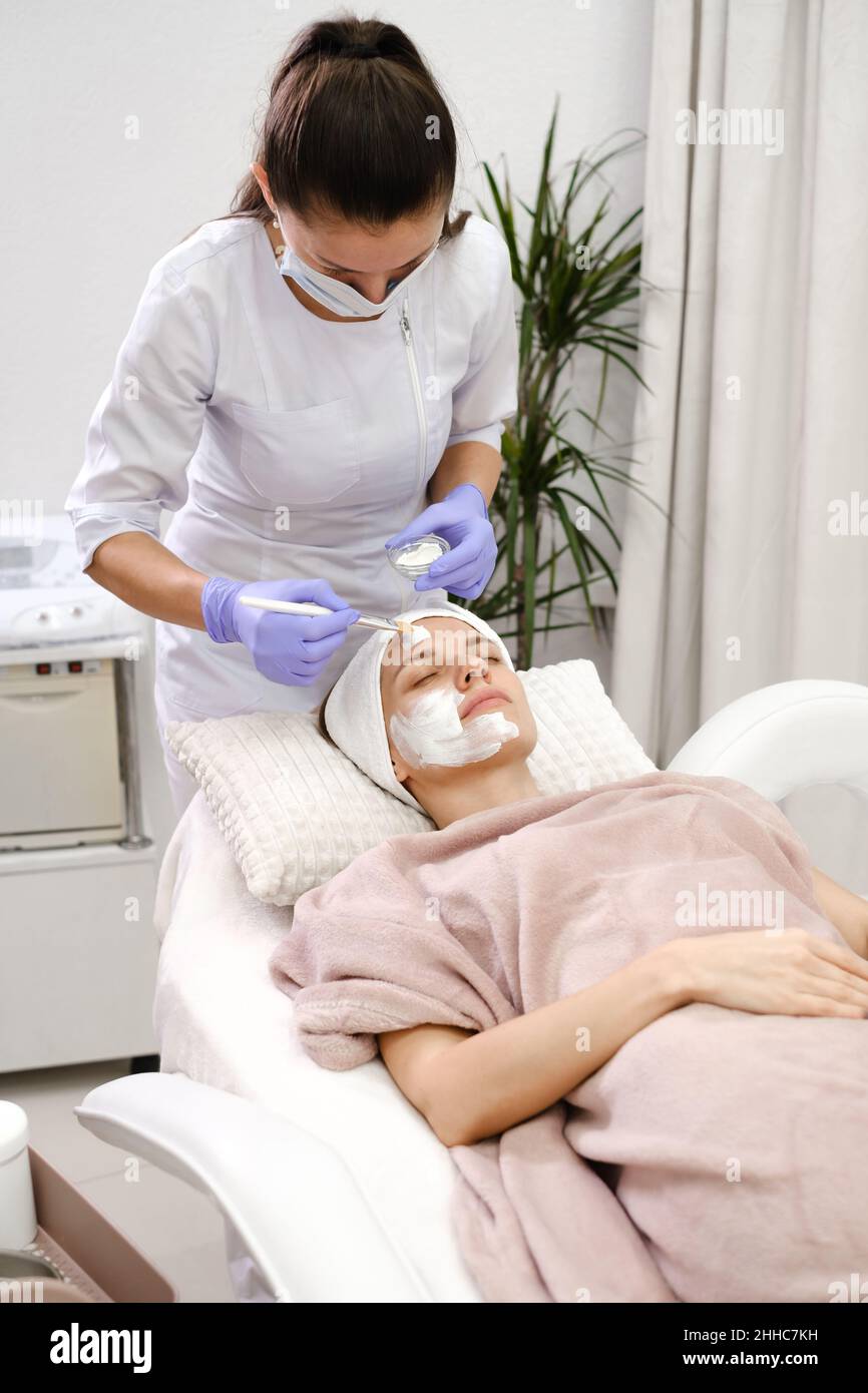 Il cosmetologo applica la maschera della pelle con pennello, cura e procedura di trattamento in clinica estetica moderna. Donna tratta la pelle in salone di bellezza, anti-età non Foto Stock
