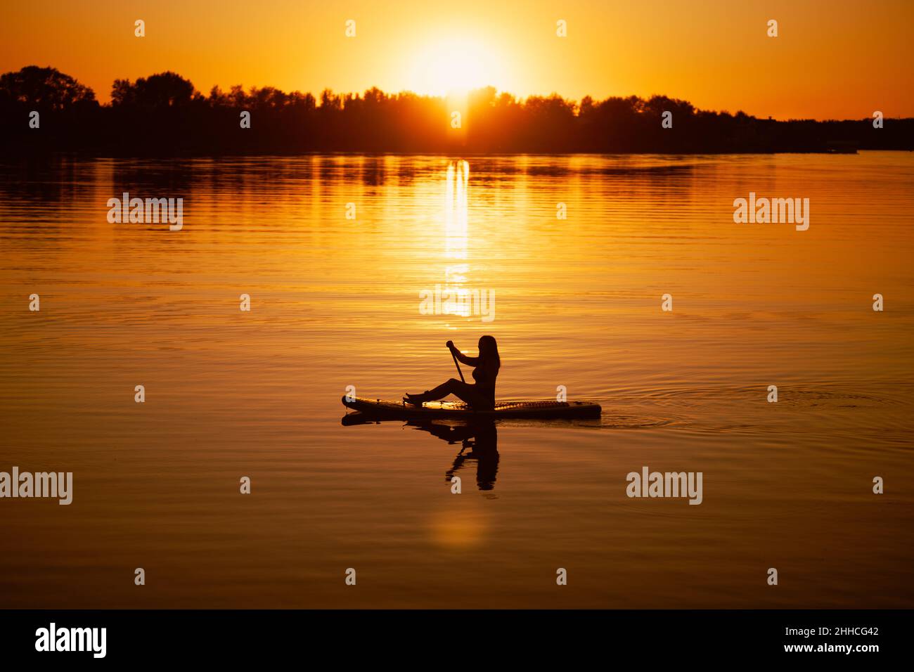 Paddle boarding signora silhouette con il remo in mano sul lago con incantevole tramonto sullo sfondo che copre l'acqua di colore arancione in estate. Attivo Foto Stock