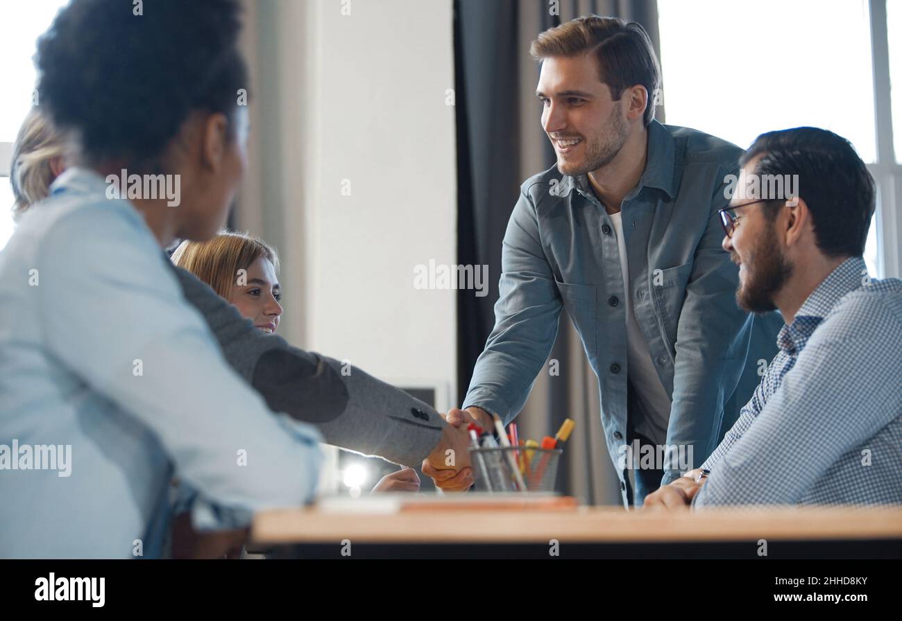 Ottimo lavoro. Due uomini d'affari allegri scuotono le mani mentre i loro colleghi applaudono e sorridono sullo sfondo Foto Stock