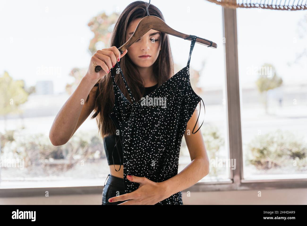 Giovane bruna che tocca e ispeziona l'abito punteggiato alla moda mentre ti trovi vicino alla finestra durante il giorno a casa Foto Stock