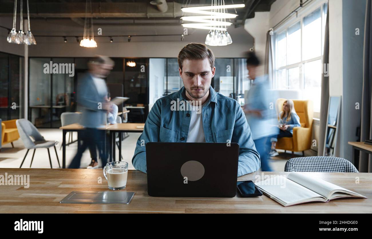 Un dipendente freelance di giovani millenni che lavora al progetto in un notebook durante la corsa in ufficio in una grande e moderna sala di lavoro Foto Stock