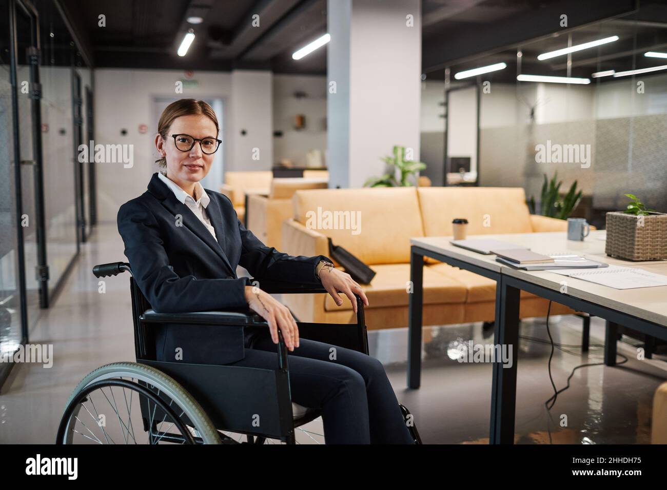 Ritratto di una donna d'affari di successo con sedia a rotelle e guardando la fotocamera sorridendo mentre si posa in ufficio moderno spazio Foto Stock