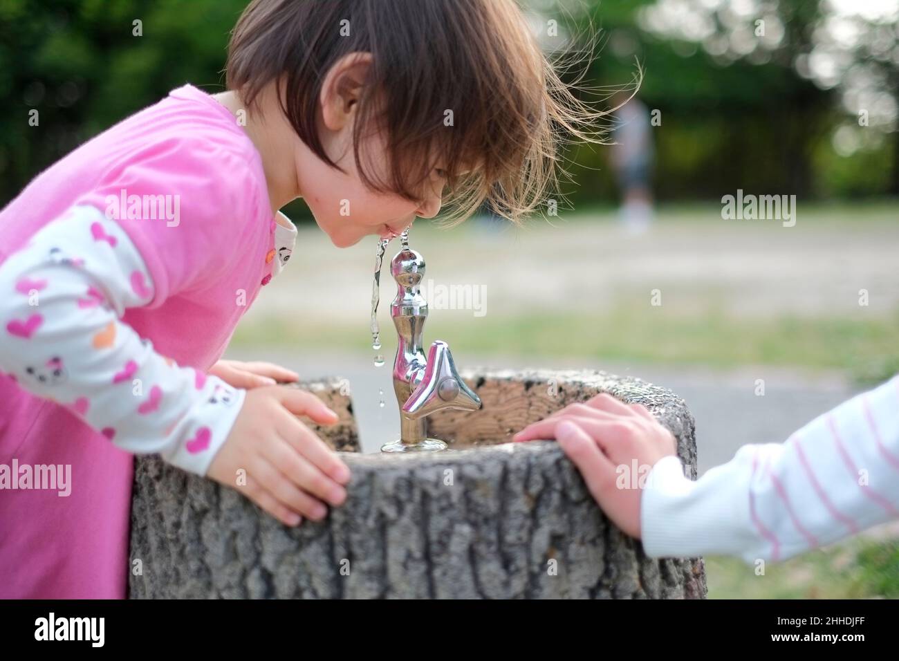 Bambina con capelli corti marroni che bevono acqua da una fontana nel parco. Primavera. Foto Stock